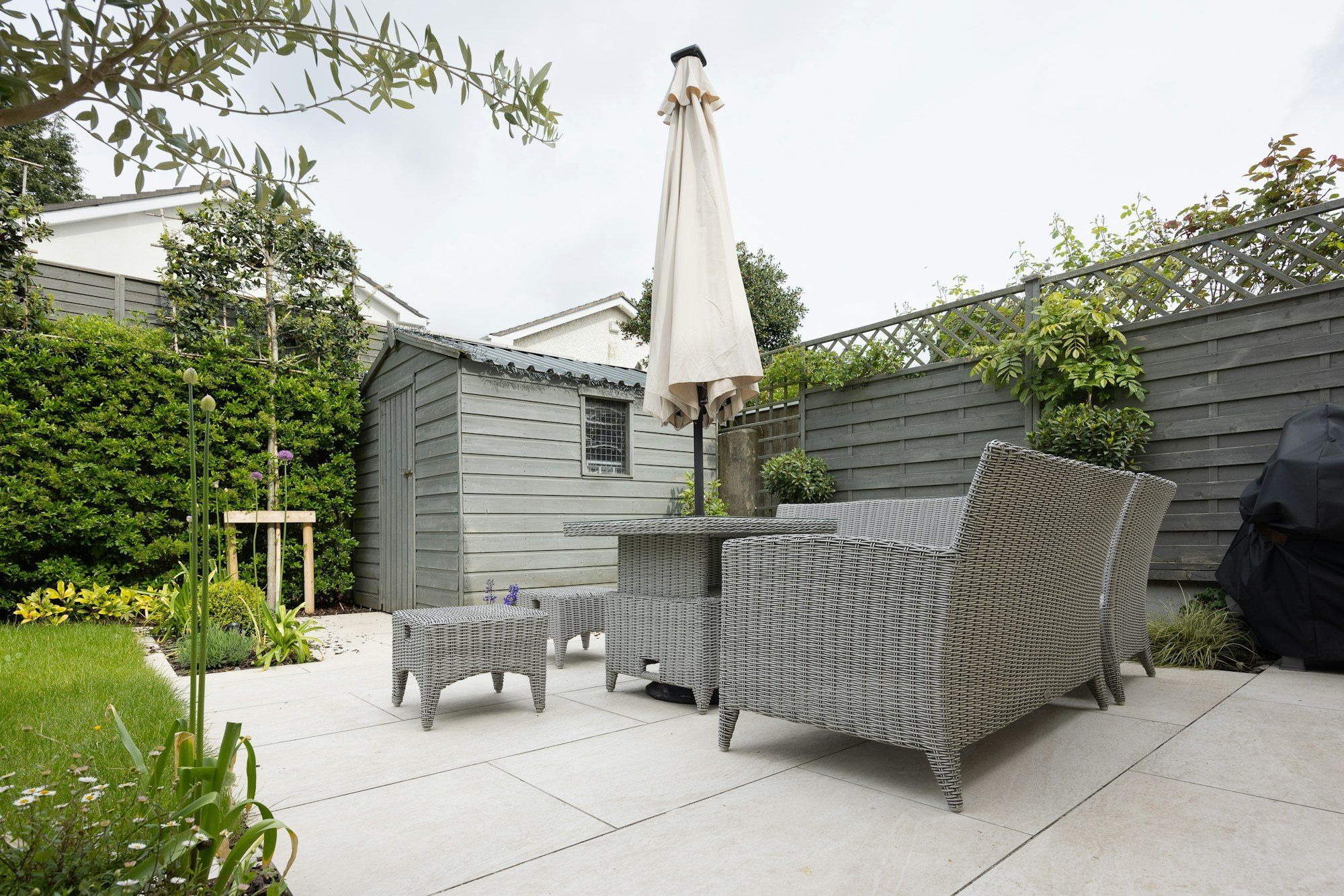 Pale paved patio with a parasol, seating and a garden shed